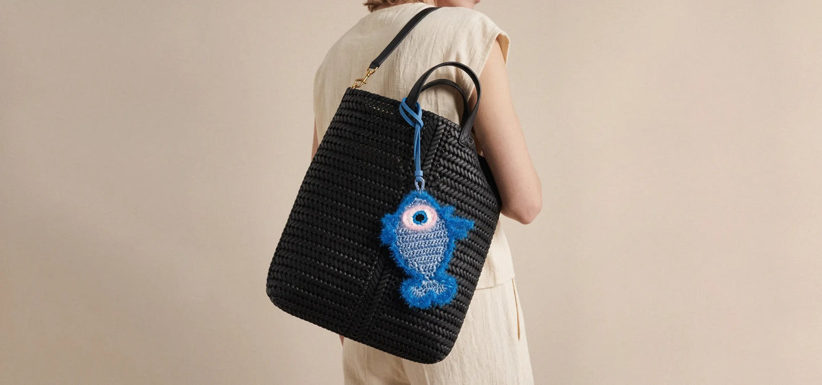 A person carries a black woven tote bag with a blue woven fish charm hanging from the handle