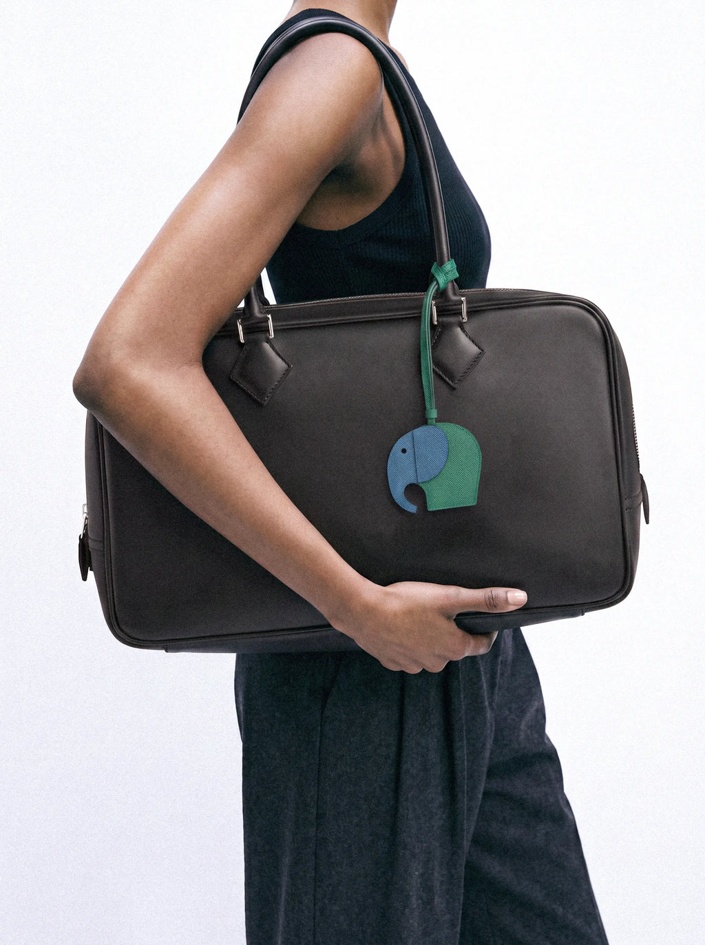 A person in a black sleeveless top and dark pants carries a large black leather duffel bag with a green and blue elephant charm hanging from the zipper.