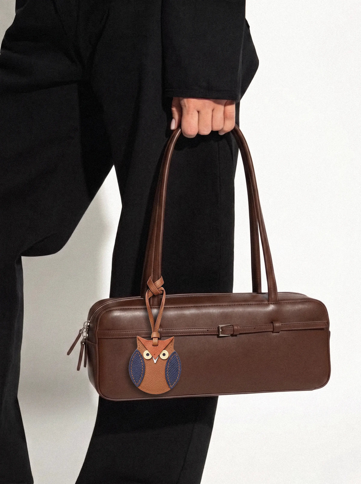A person in a black outfit holds a brown leather duffel-style bag with long handles and an owl charm on the front. The owl has blue wings and a white eye patch, set against a plain light background.