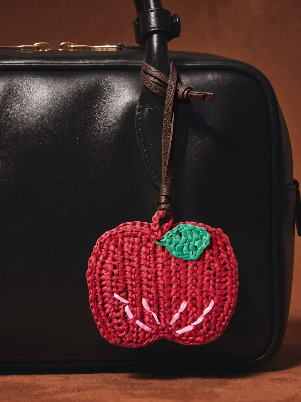 A red crocheted apple bag charm with a green leaf and white stitched details, hanging from a brown leather strap on a black leather bag against a warm brown background.