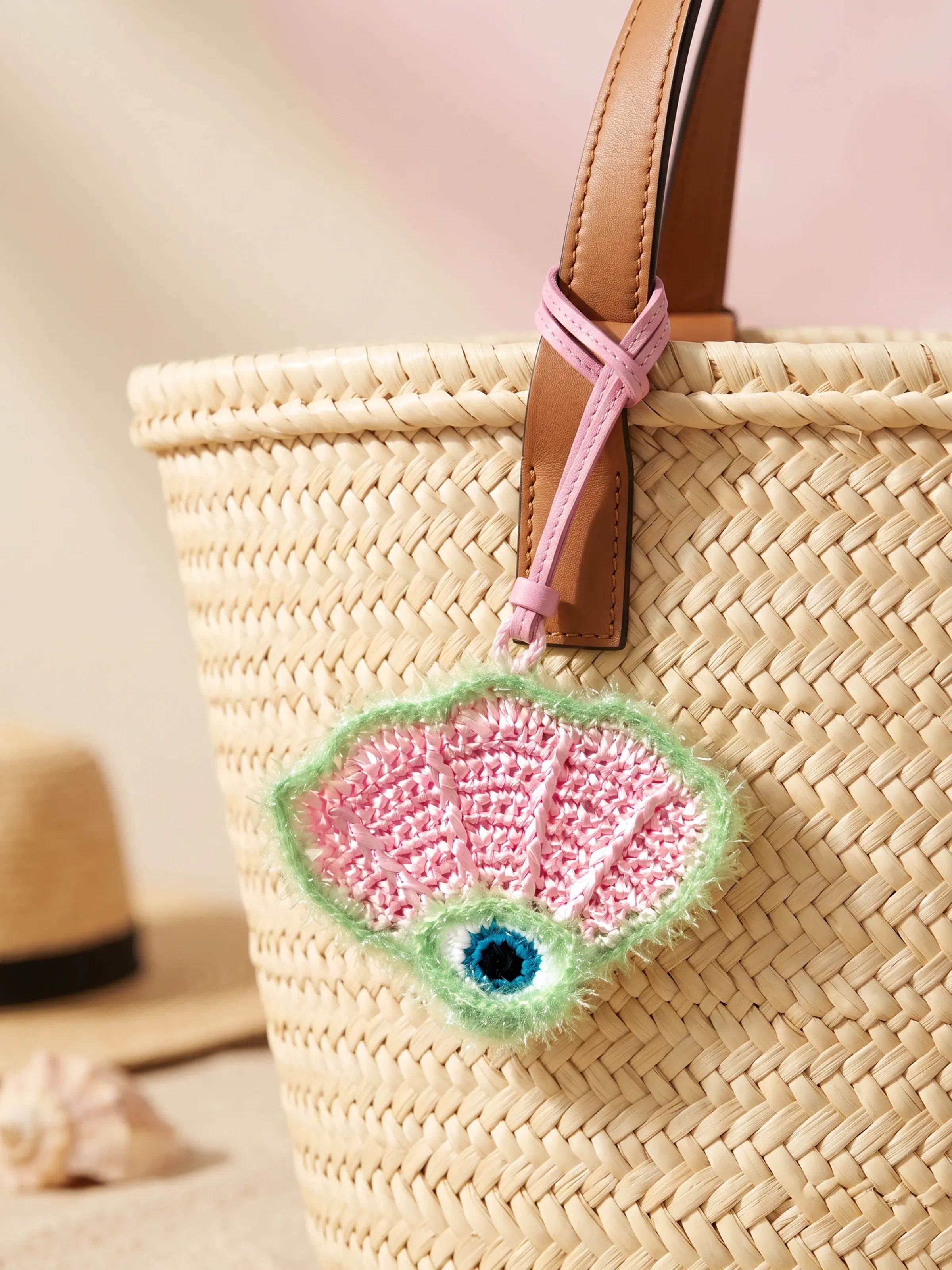 A woven straw tote bag with a tan leather handle, decorated with a dangling shell-shaped charm in pink, green, and blue.