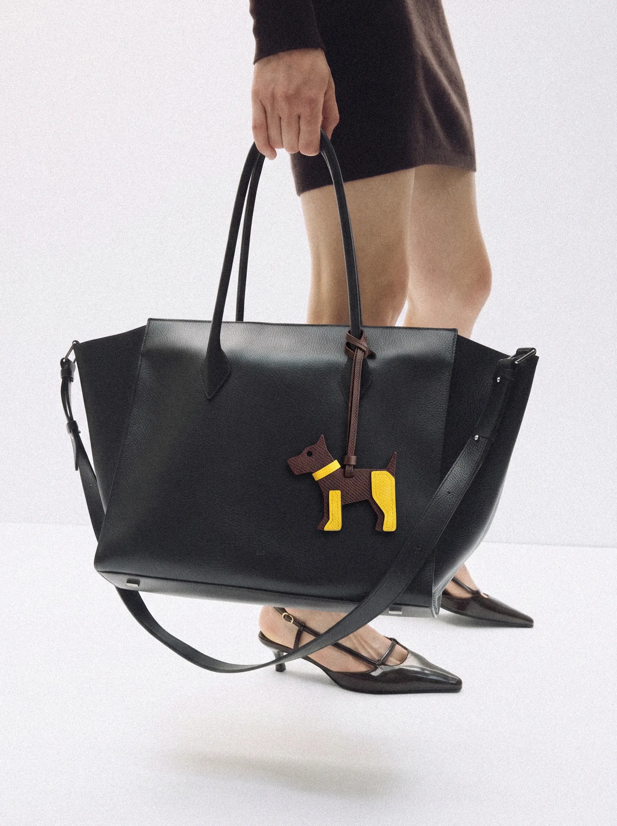 A model holds a large black leather tote bag featuring a sculptural brown and yellow dog charm. 