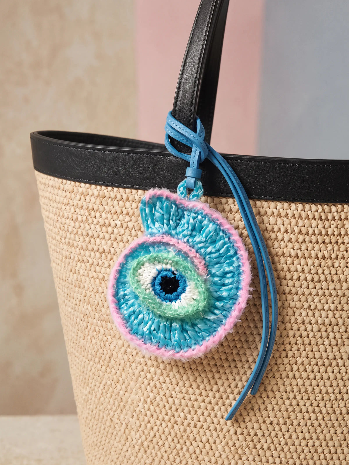 A woven straw tote bag with a colorful crocheted evil eye charm hanging from the handle.