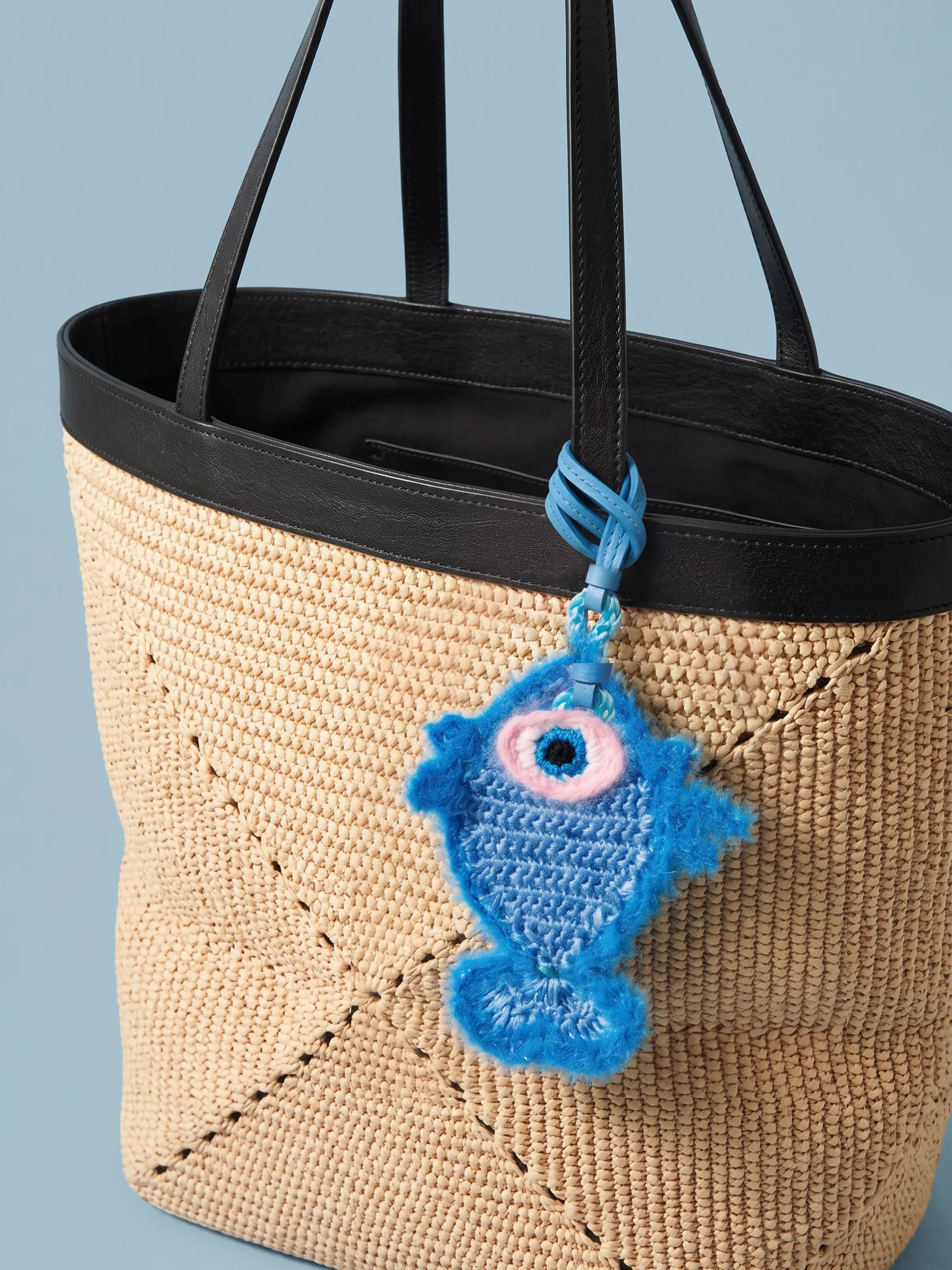 A woven straw tote bag with black trim and handles, featuring a blue fish charm with a pink eye hanging from one handle, set against a solid light blue background.