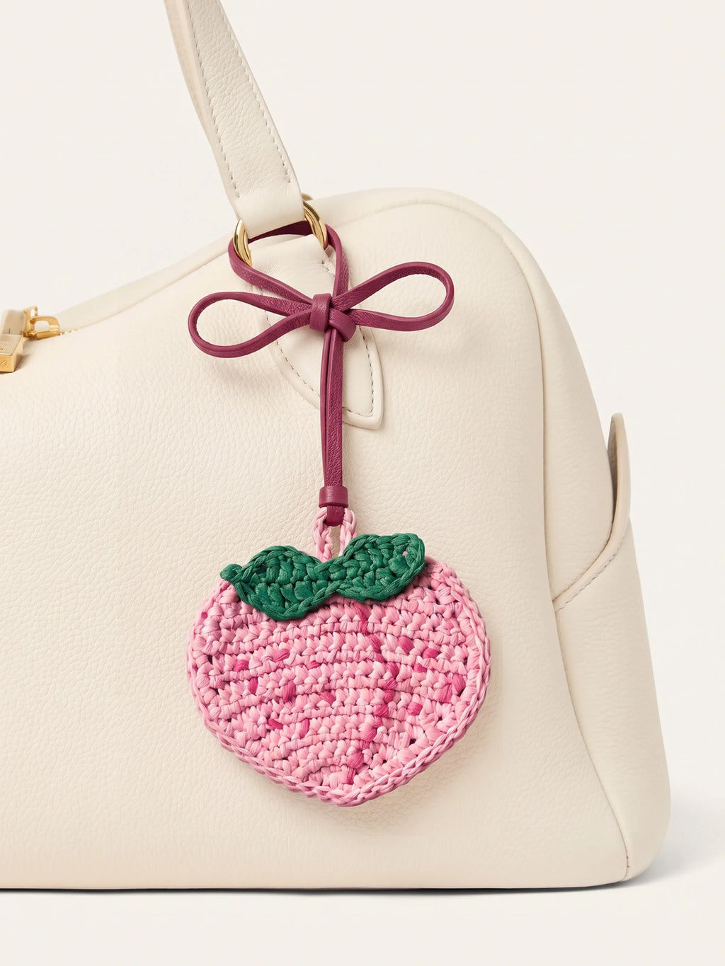 A cream-colored leather handbag featuring a pink crocheted peach charm with green leaves and a burgundy ribbon bow, attached to the side.