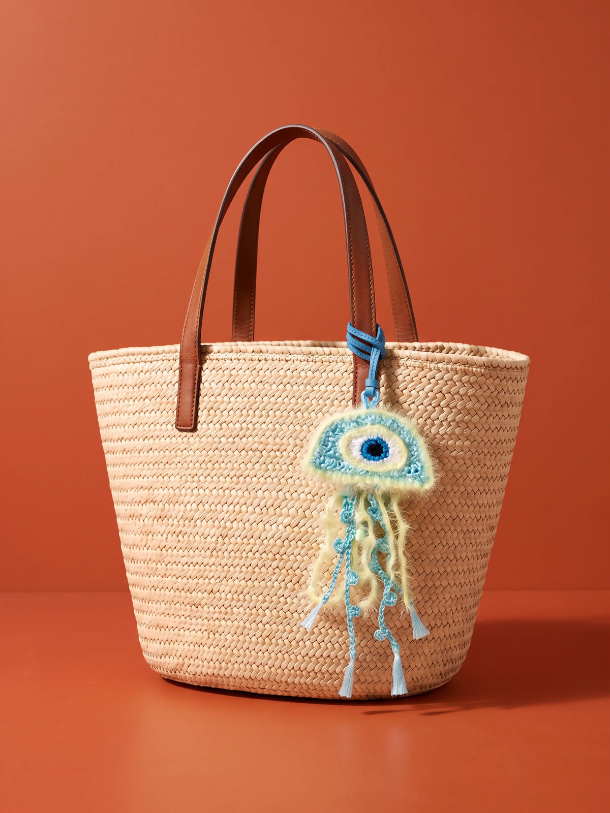 A woven straw tote bag with brown leather handles, featuring a hanging blue and white crocheted jellyfish charm, set against a solid terracotta orange background.