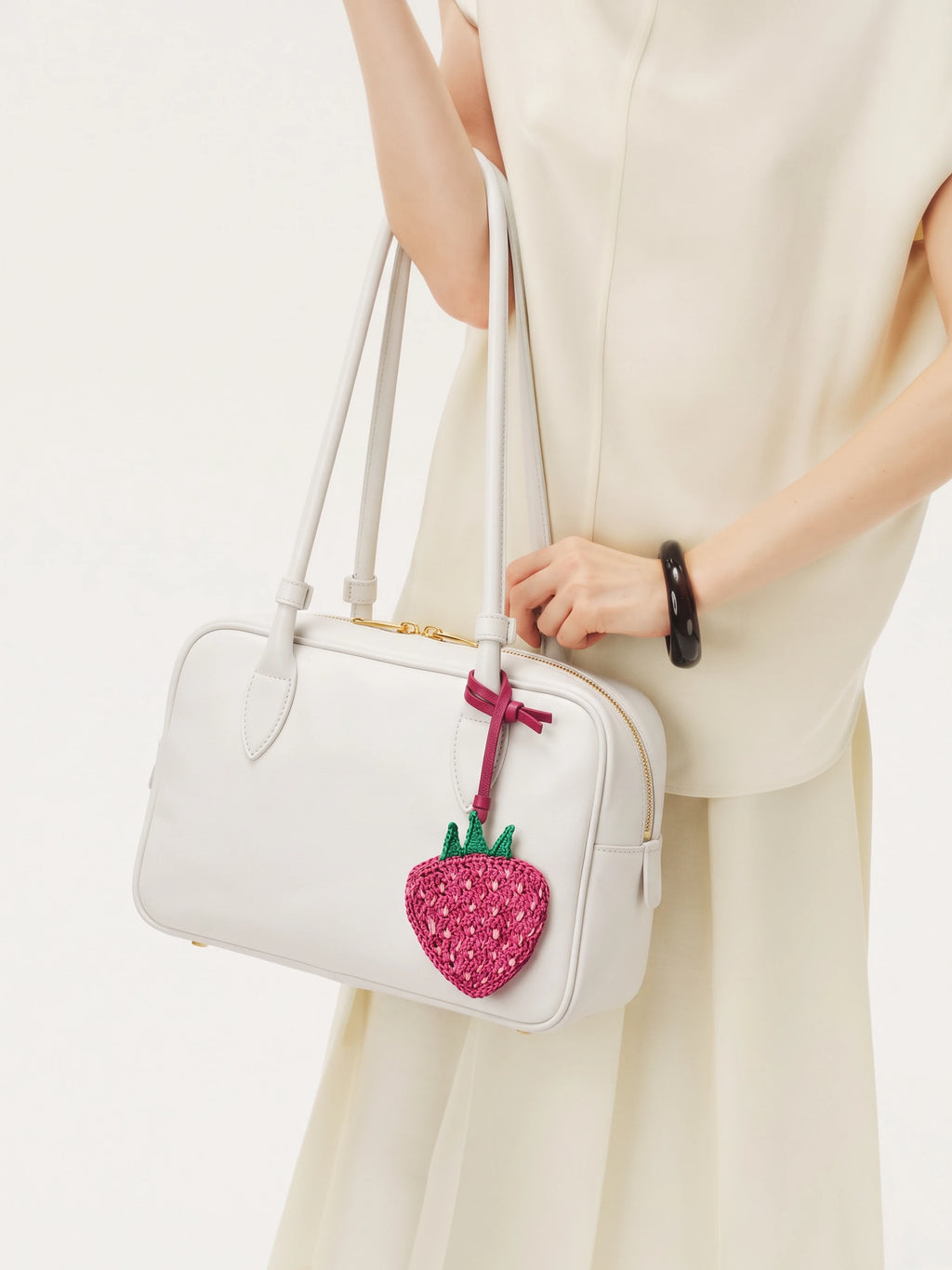 A white structured handbag with double shoulder straps and a pink strawberry charm, carried by a model wearing a cream-colored sleeveless dress.