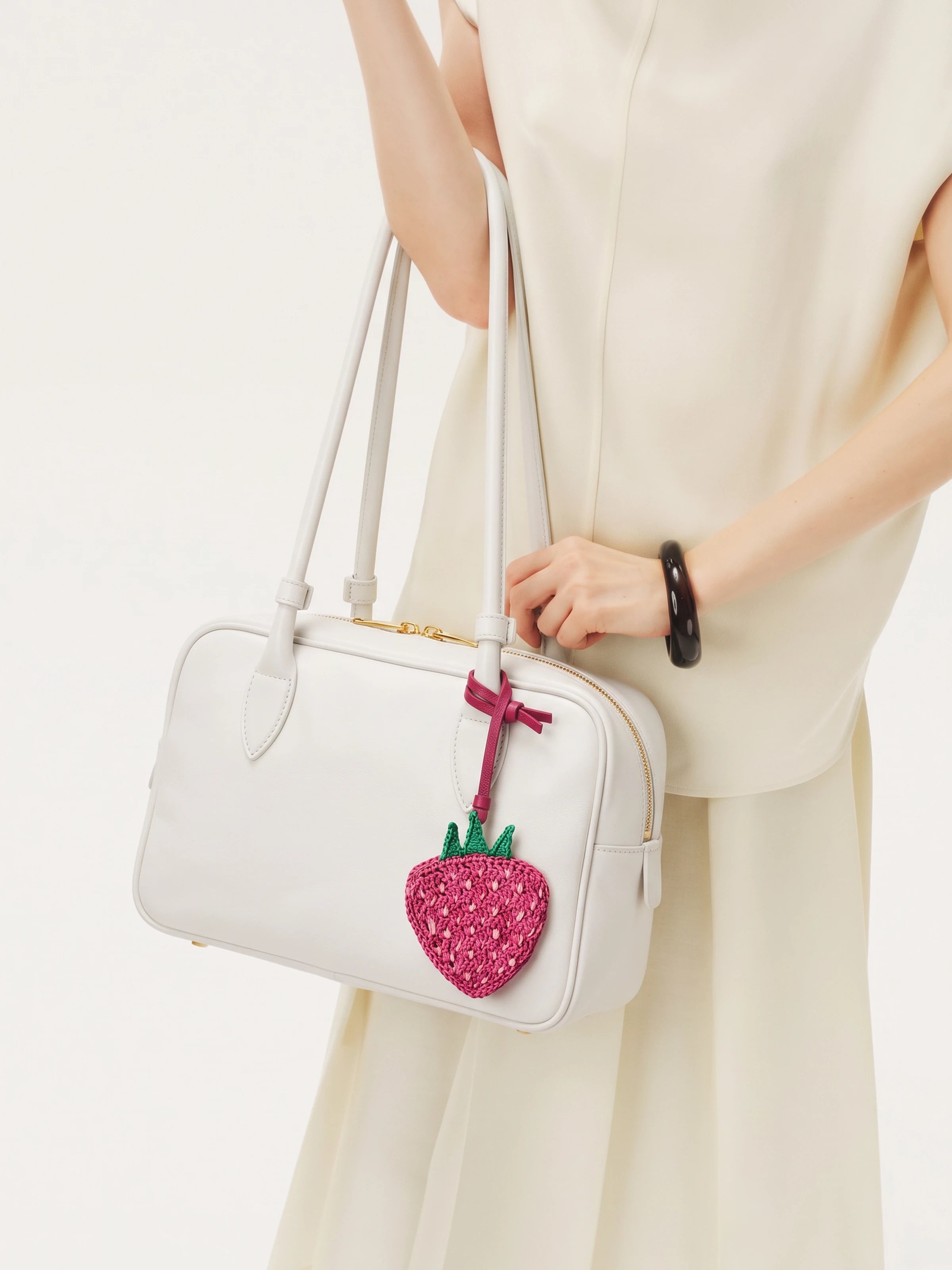 A white structured handbag with double shoulder straps and a pink strawberry charm, carried by a model wearing a cream-colored sleeveless dress.