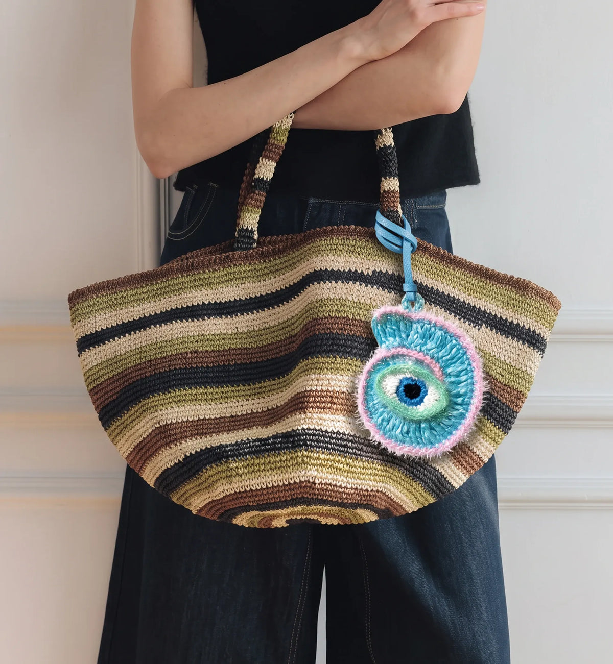 A person holds a woven bag with a decorative hanging charm shaped like a colorful eye with blue, green, pink, and white details.
