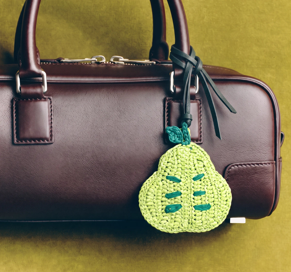 A dark brown leather handbag with a green raffia pear-shaped charm