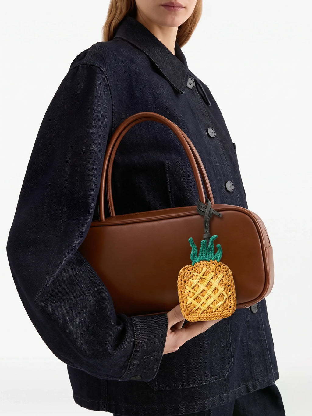 A model wearing a dark denim jacket holds a brown leather handbag with a woven pineapple-shaped charm hanging from the handle.