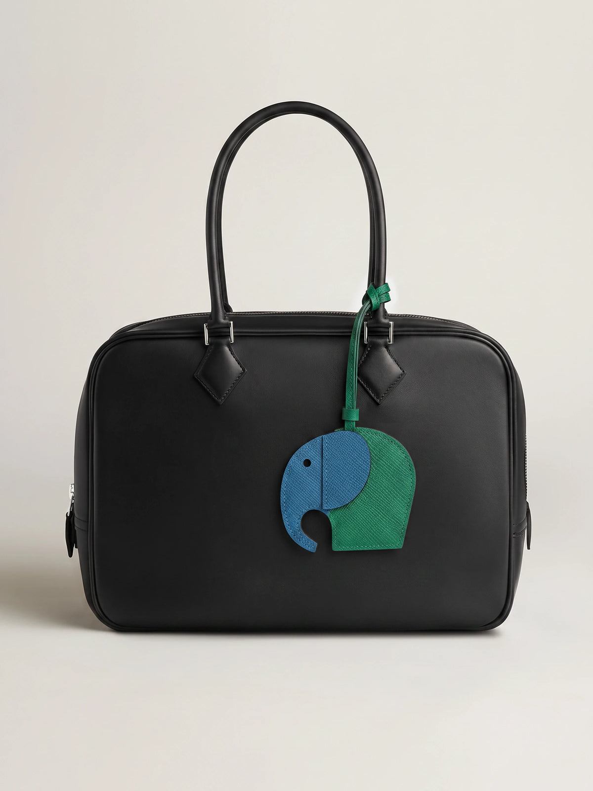 A black leather handbag with a top handle and a hanging elephant charm in blue and green, photographed against a plain light beige studio background.