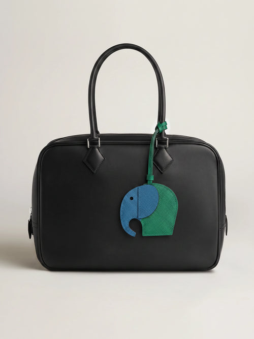 A black leather handbag with a top handle and a hanging elephant charm in blue and green, photographed against a plain light beige studio background.