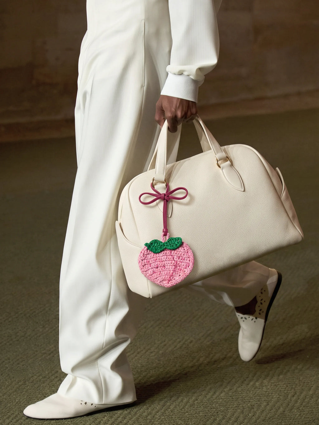 A person in an all-white outfit carries a cream-colored leather duffel bag with a pink crocheted peach charm featuring green leaves.