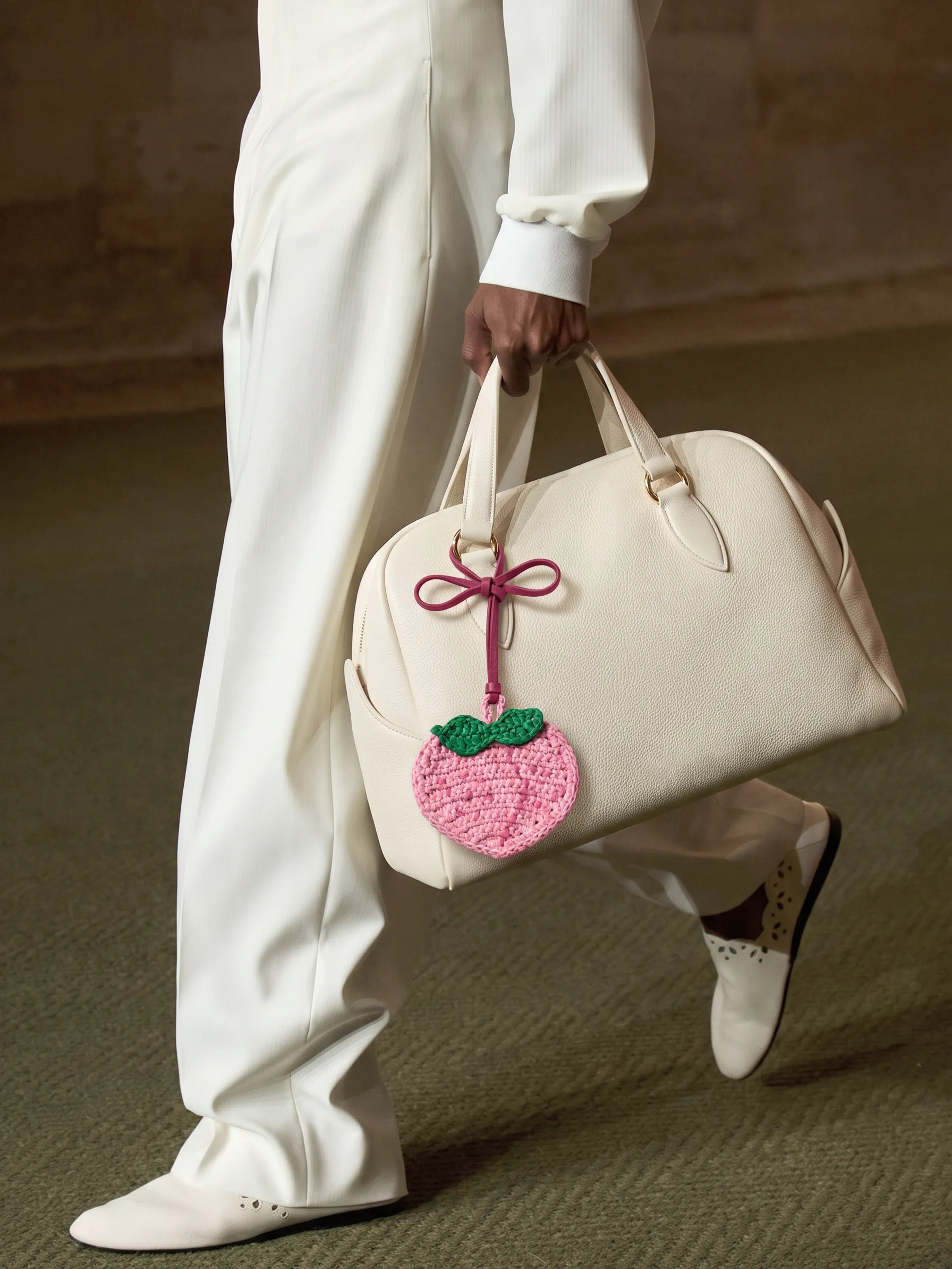 A person in an all-white outfit carries a cream-colored leather duffel bag with a pink crocheted peach charm featuring green leaves.