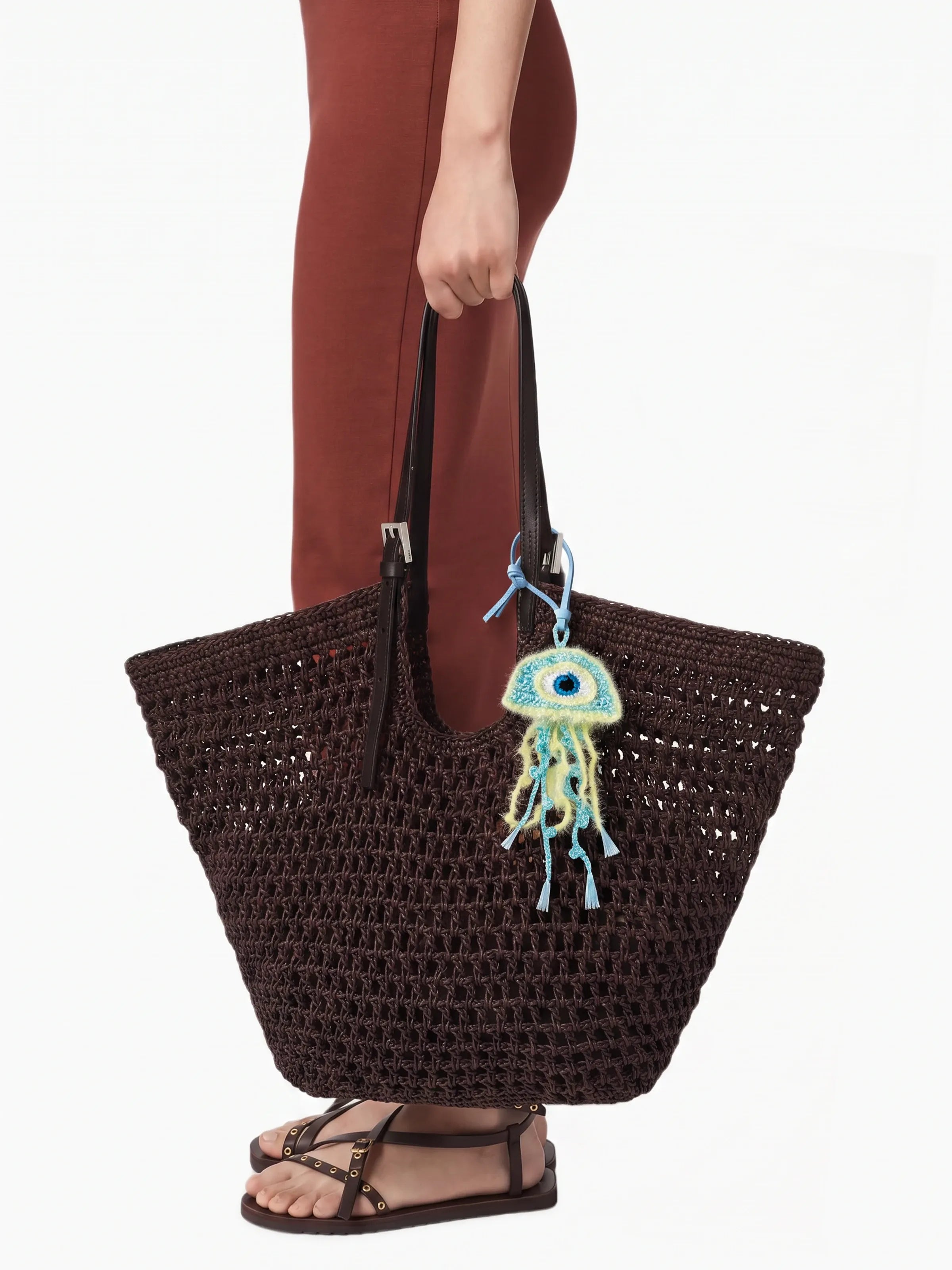A person wearing rust-brown pants and black sandals holds a large dark brown crocheted tote bag with black handles. A colorful embroidered jellyfish charm hangs from the bag.