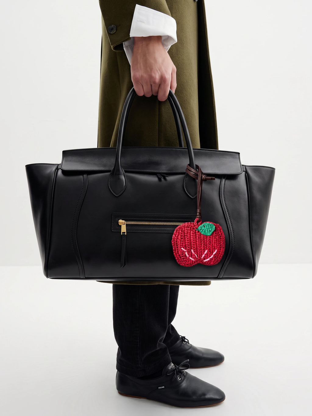 A person holds a large black leather tote bag with gold-tone hardware and a red apple hanging charm. The person is wearing an olive green coat, a white shirt, black trousers, and black shoes against a clean white studio background.
