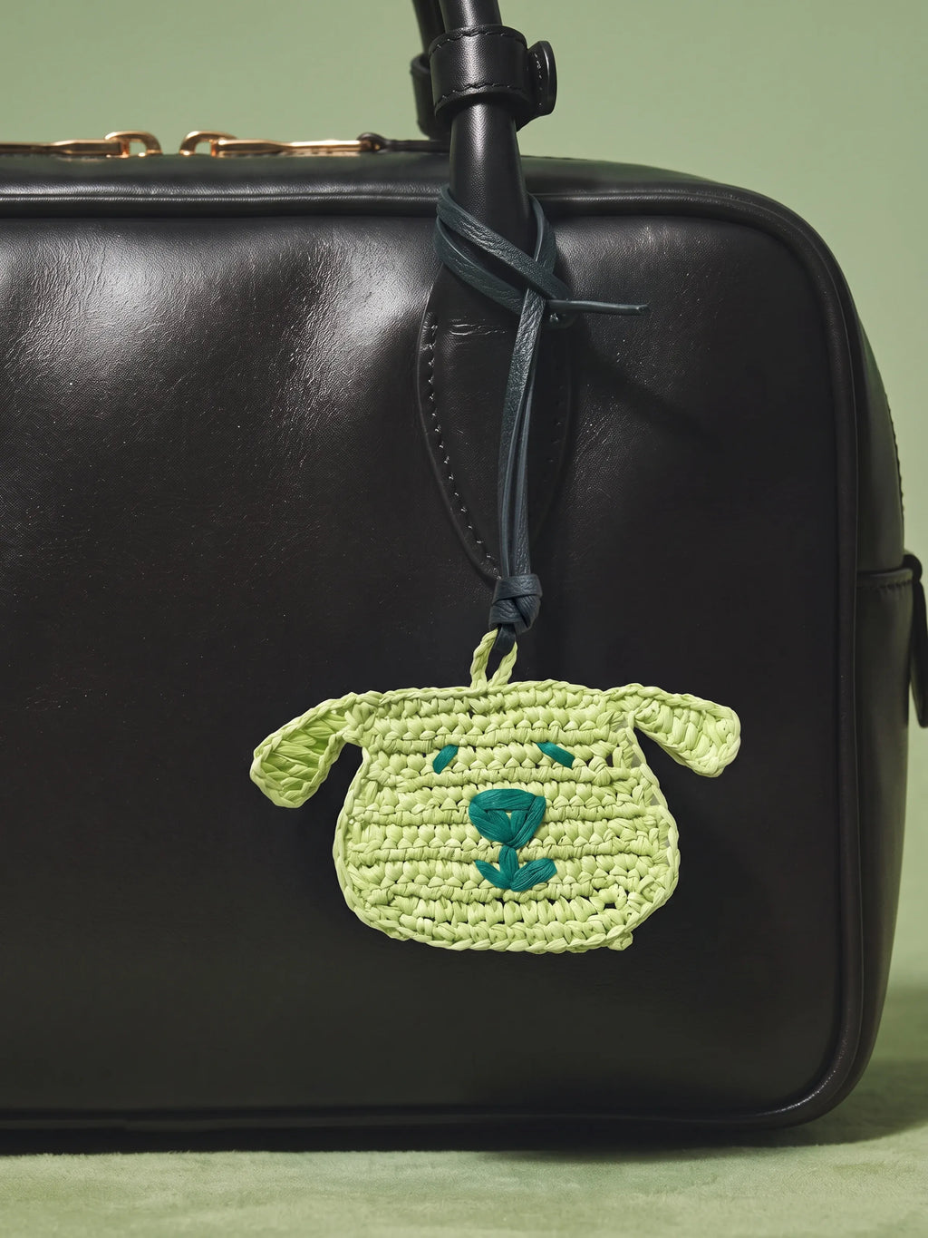 A light green crocheted dog bag charm with a teal embroidered nose hangs from a black leather bag strap against a soft sage green background.