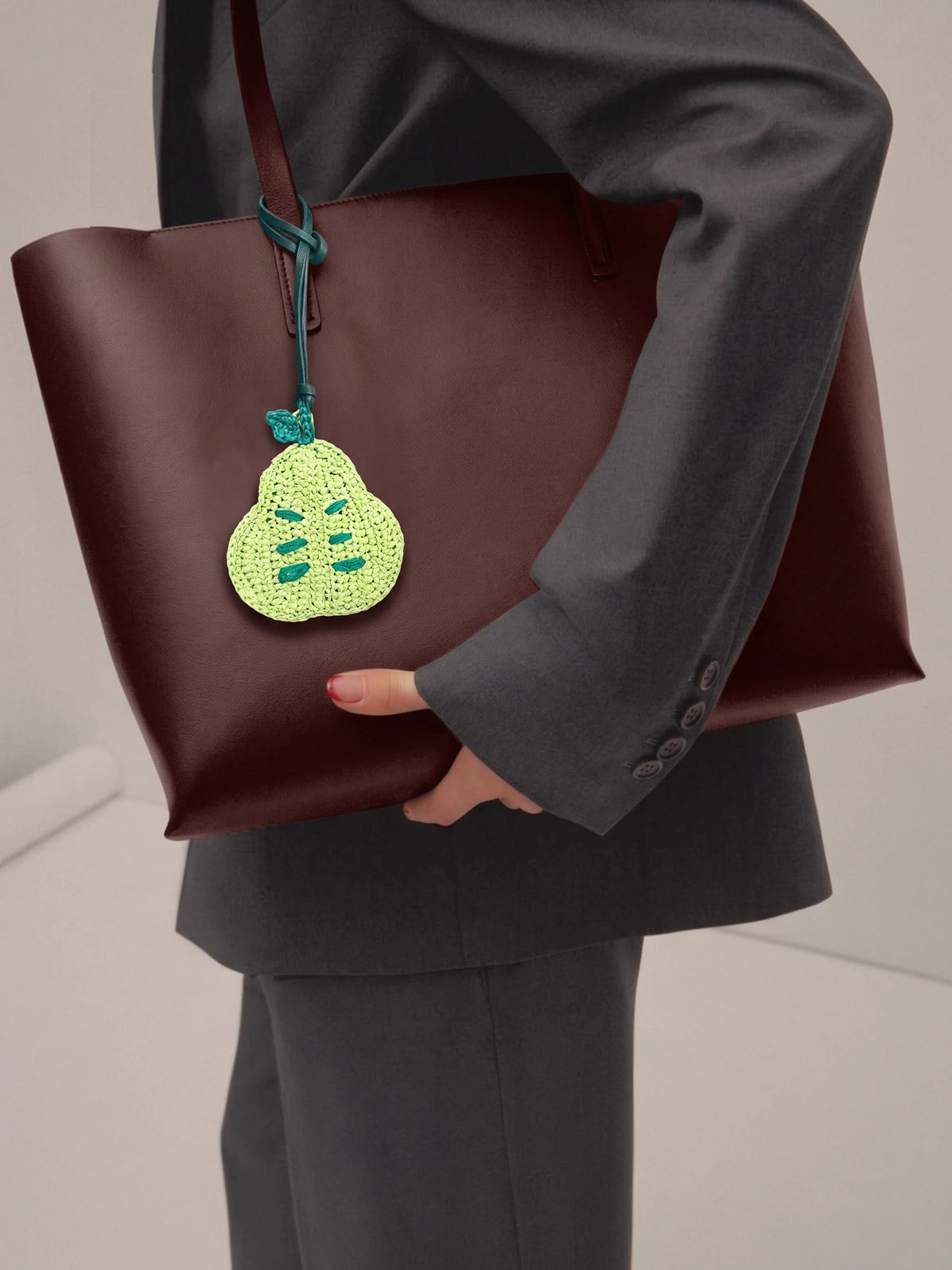 A person in a dark gray suit holding a large dark brown leather tote bag with a green crocheted pear-shaped charm hanging from the strap.