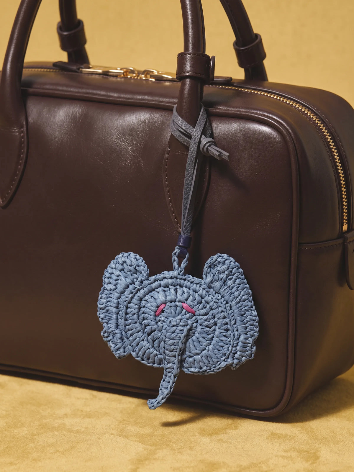 A dark brown leather handbag with a crocheted blue elephant charm hanging from its zipper, set against a warm beige background.
