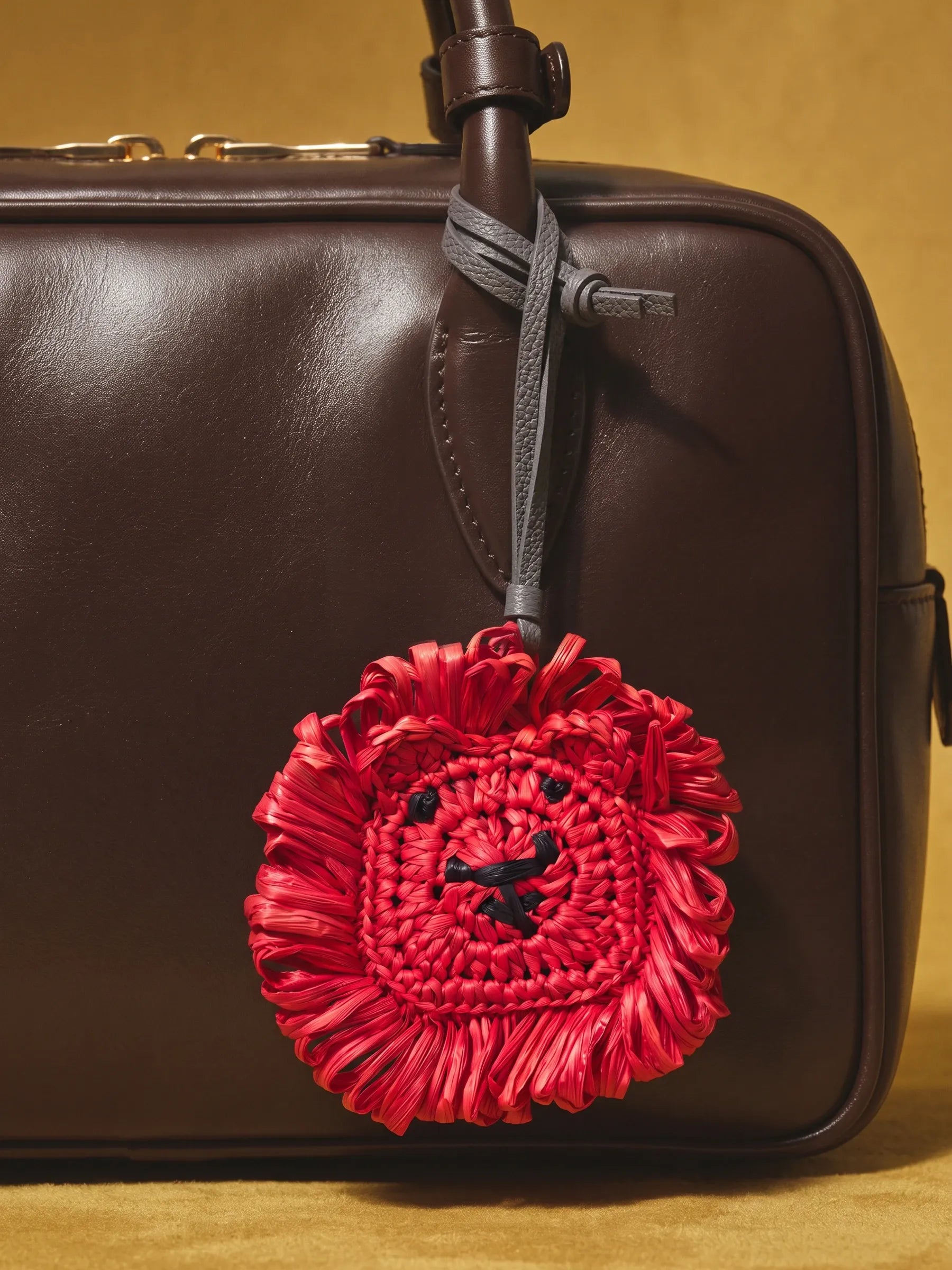 A close-up of a dark brown leather bag with a hanging red woven lion-face charm and gray strap, set against a warm tan background.