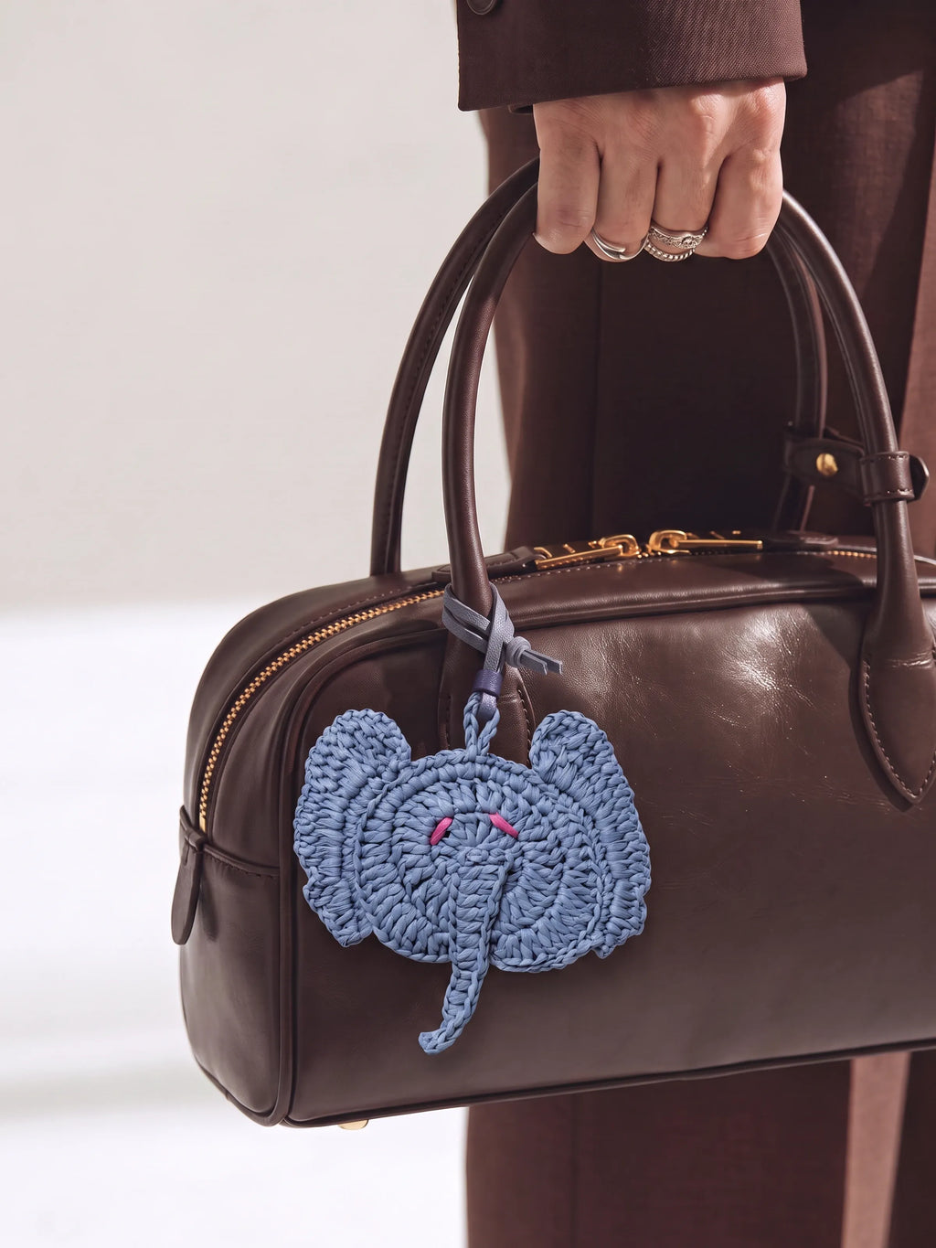 A dark brown leather handbag with gold-tone hardware, held by a person wearing brown clothing and silver rings. A light blue crocheted elephant charm hangs from the bag handle.