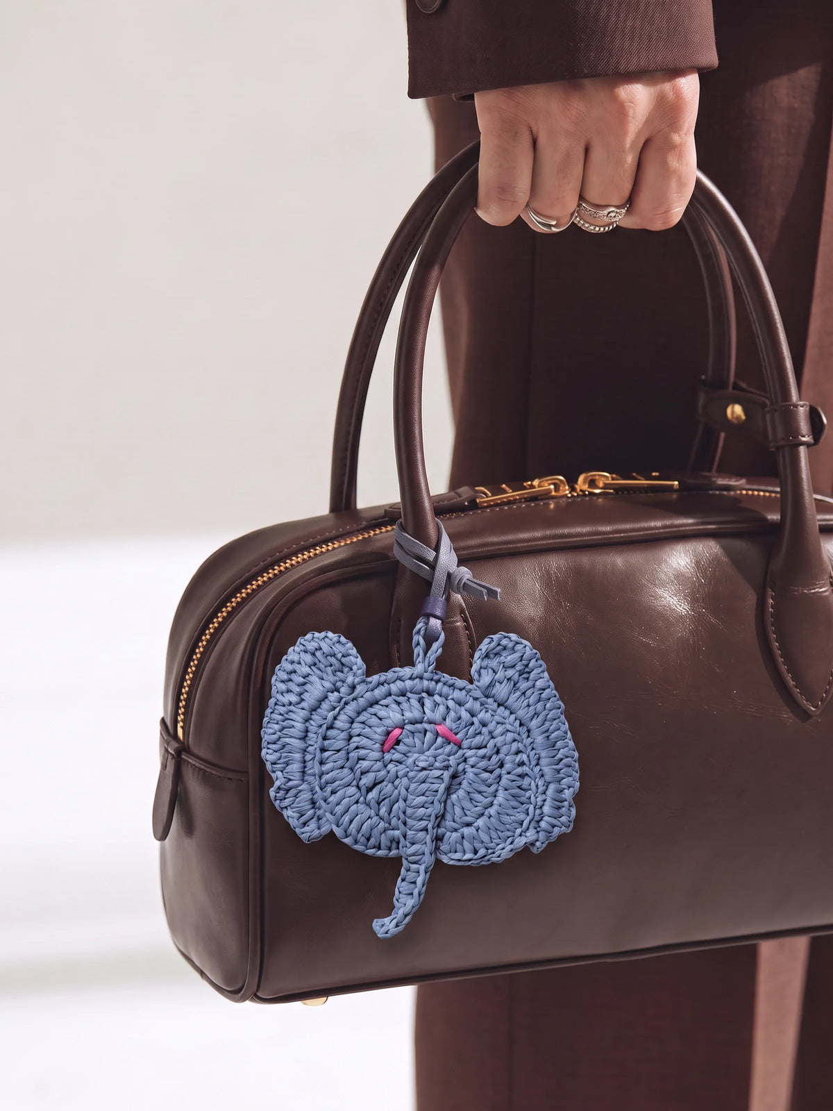 A dark brown leather handbag with gold-tone hardware, held by a person wearing brown clothing and silver rings. A light blue crocheted elephant charm hangs from the bag handle.