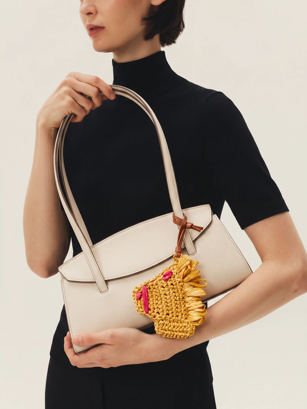 A model in a black turtleneck sweater holds a structured cream-colored leather handbag with a long handle and a yellow woven horse charm with pink accents.