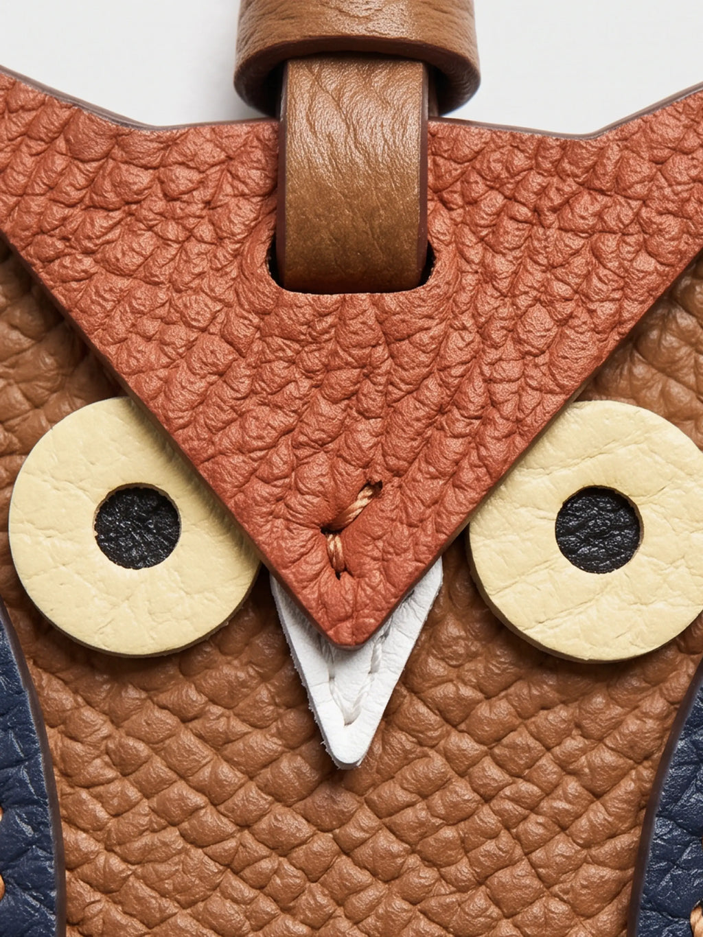 Close-up of an owl-themed leather charm featuring textured brown and terracotta panels, cream-colored circular eyes, and a white beak detail.