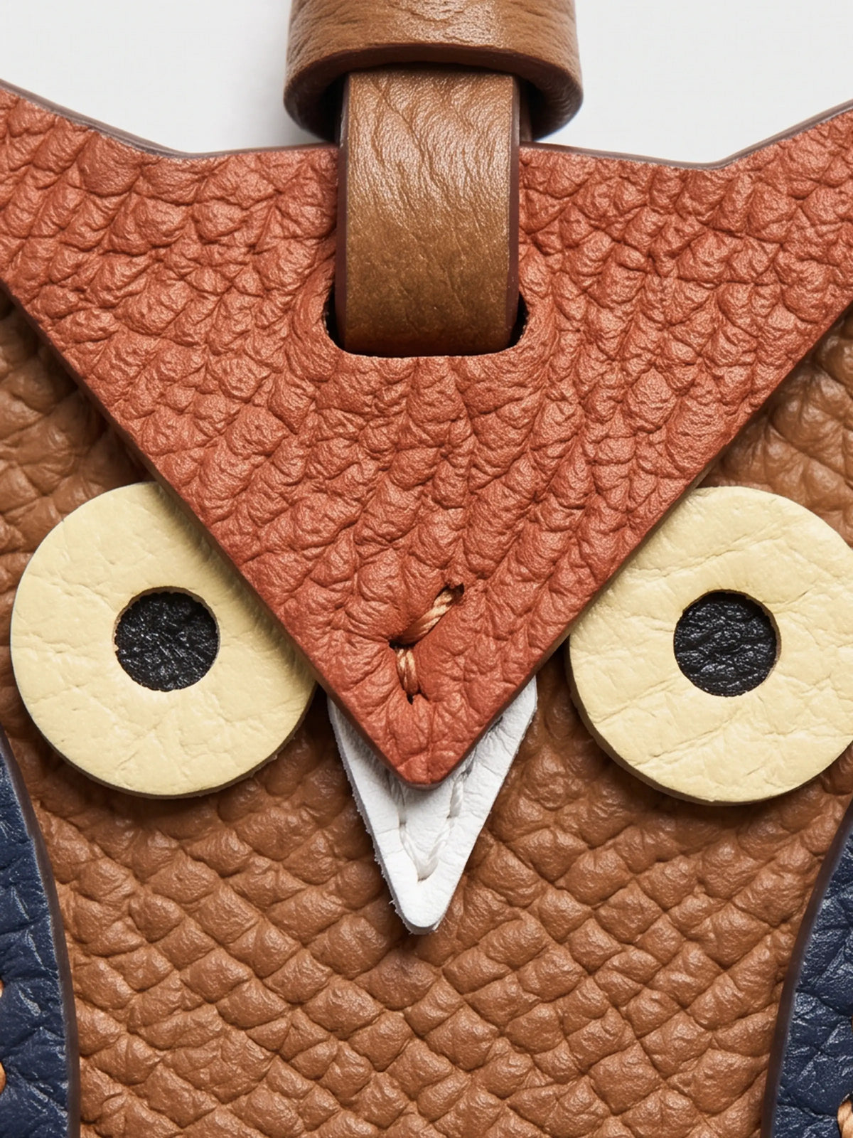 Close-up of an owl-themed leather charm featuring textured brown and terracotta panels, cream-colored circular eyes, and a white beak detail.