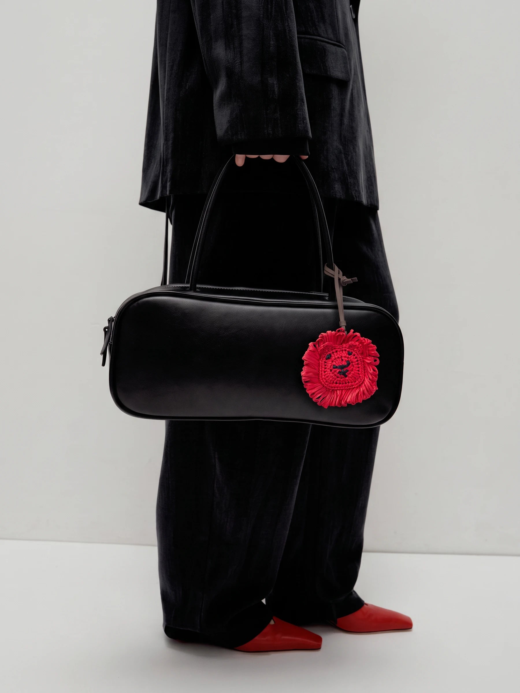 A person wearing a black velvet blazer and wide-leg trousers holds a black cylindrical handbag with a bright red lion raffia charm. The person is also wearing red pointed shoes, photographed against a plain light gray studio background.