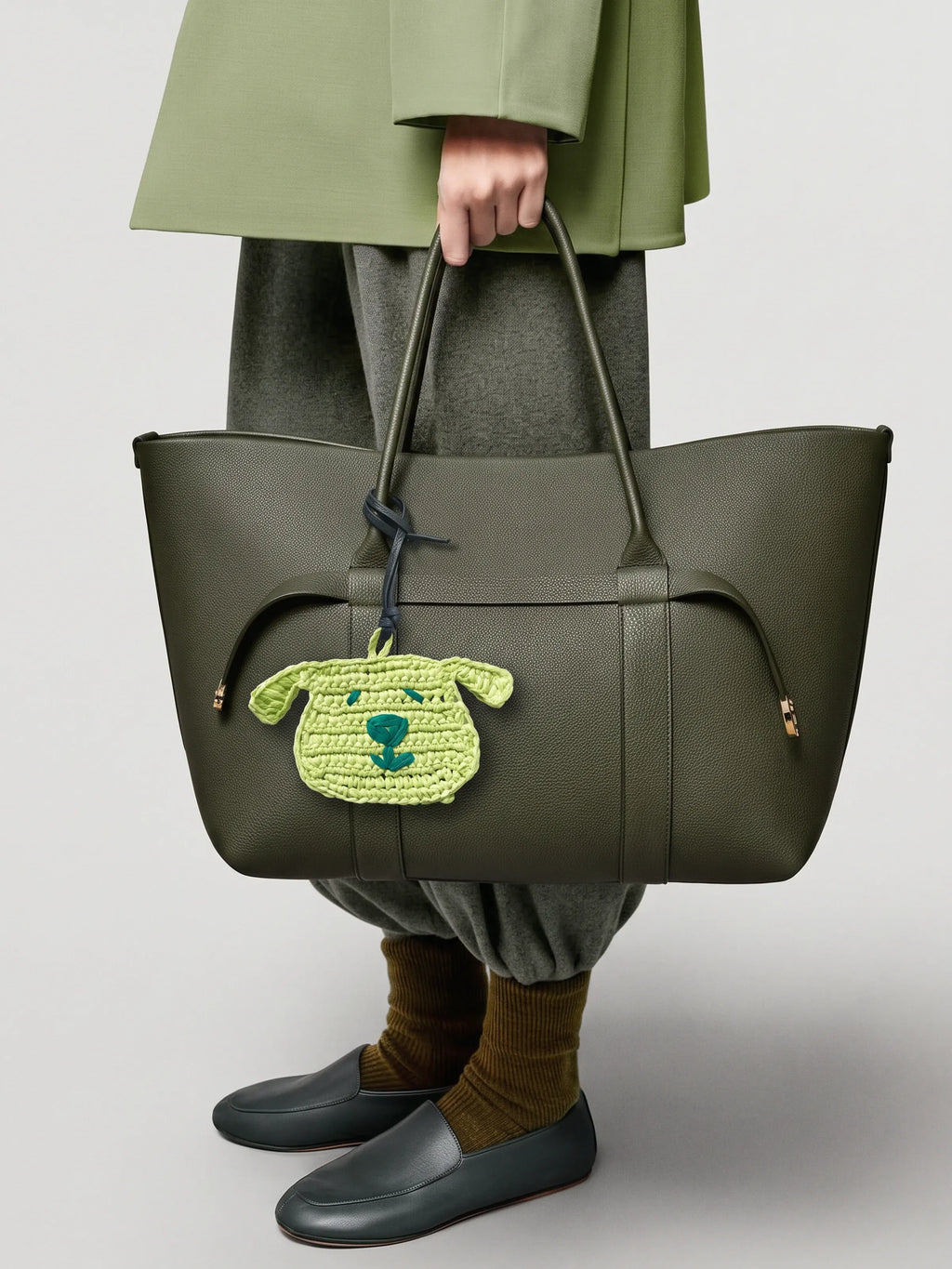 An olive green leather tote bag with a small green raffia dog charm, held by a model wearing an olive green coat, grey trousers, brown socks, and dark loafers against a light gray studio background.