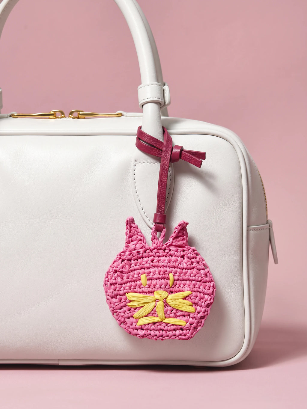 A pink crocheted cat bag charm with yellow embroidered details hangs from a burgundy ribbon attached to a white handbag, set against a solid pink background.