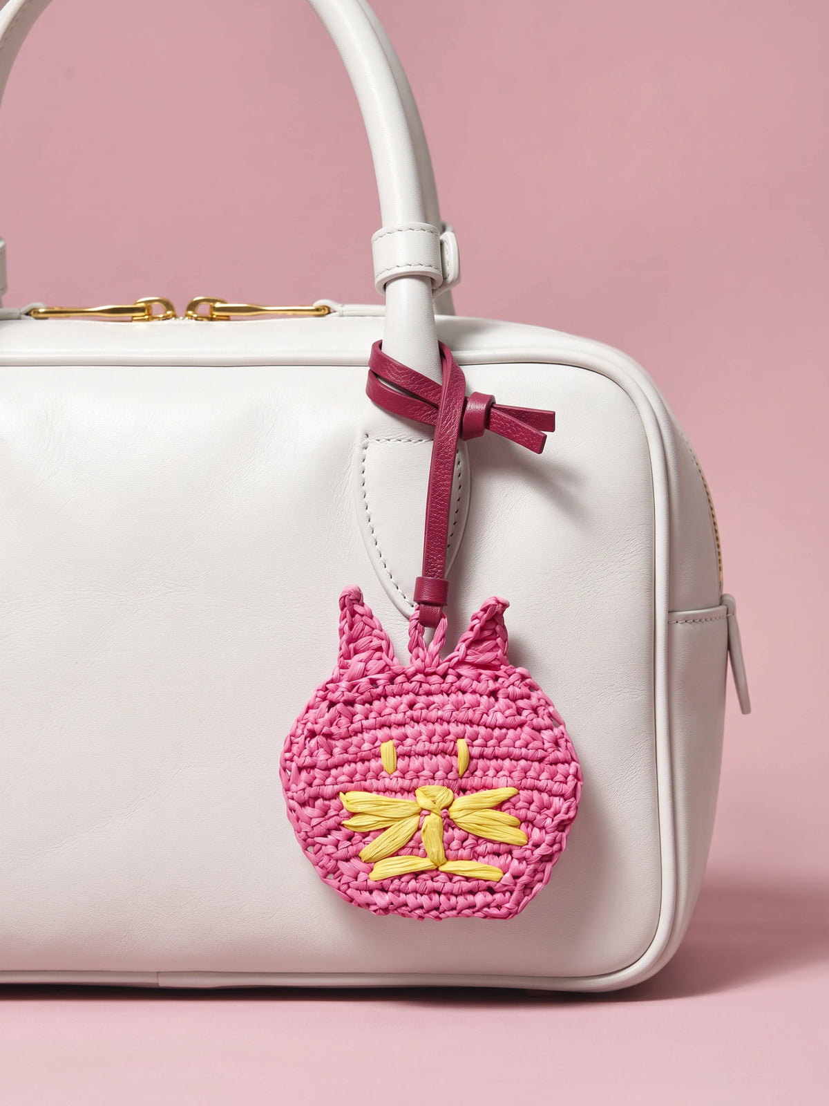 A pink crocheted cat bag charm with yellow embroidered details hangs from a burgundy ribbon attached to a white handbag, set against a solid pink background.