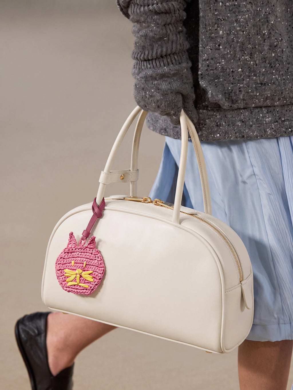A cream-colored leather handbag with a pink embroidered cat charm, carried by a person wearing a gray knit sweater and a light blue skirt.