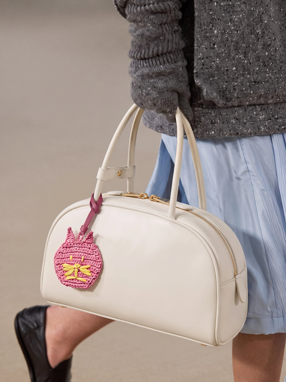 A cream-colored leather handbag with a pink embroidered cat charm, carried by a person wearing a gray knit sweater and a light blue skirt.