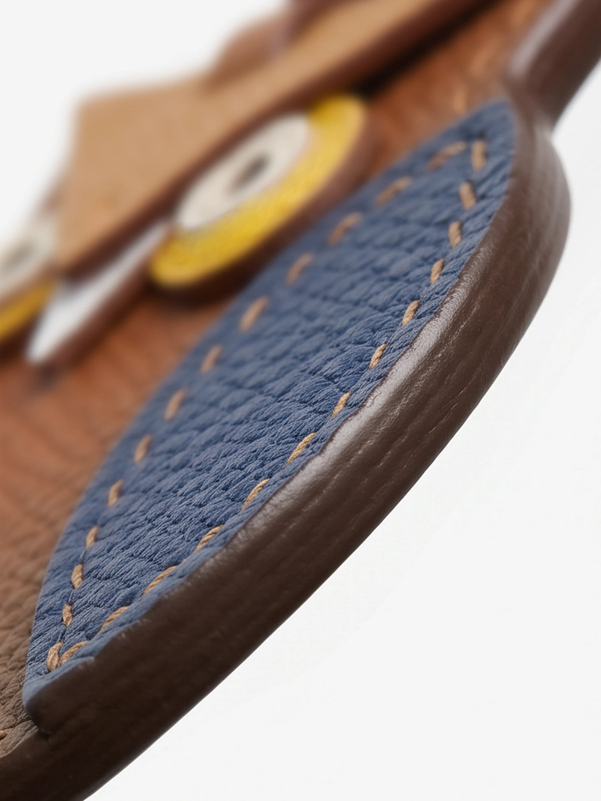 Close-up detail of a brown leather accessory with a textured blue patch and contrast stitching, showing part of a yellow circular element in the background.