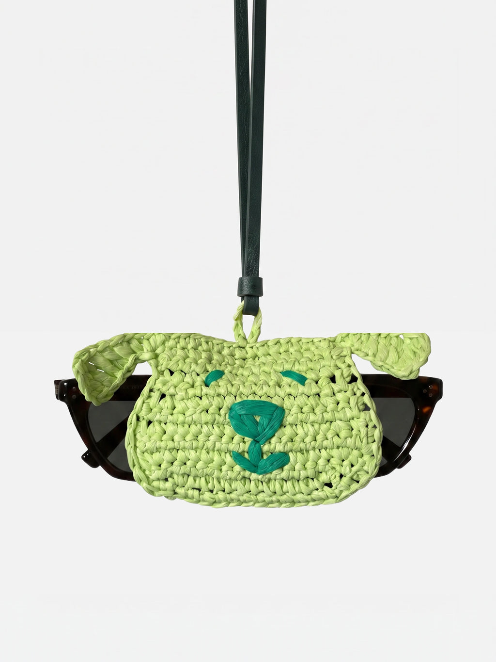 A light green woven sunglasses holder with a dog face design, holding a pair of black sunglasses, suspended from a dark strap against a light gray background.