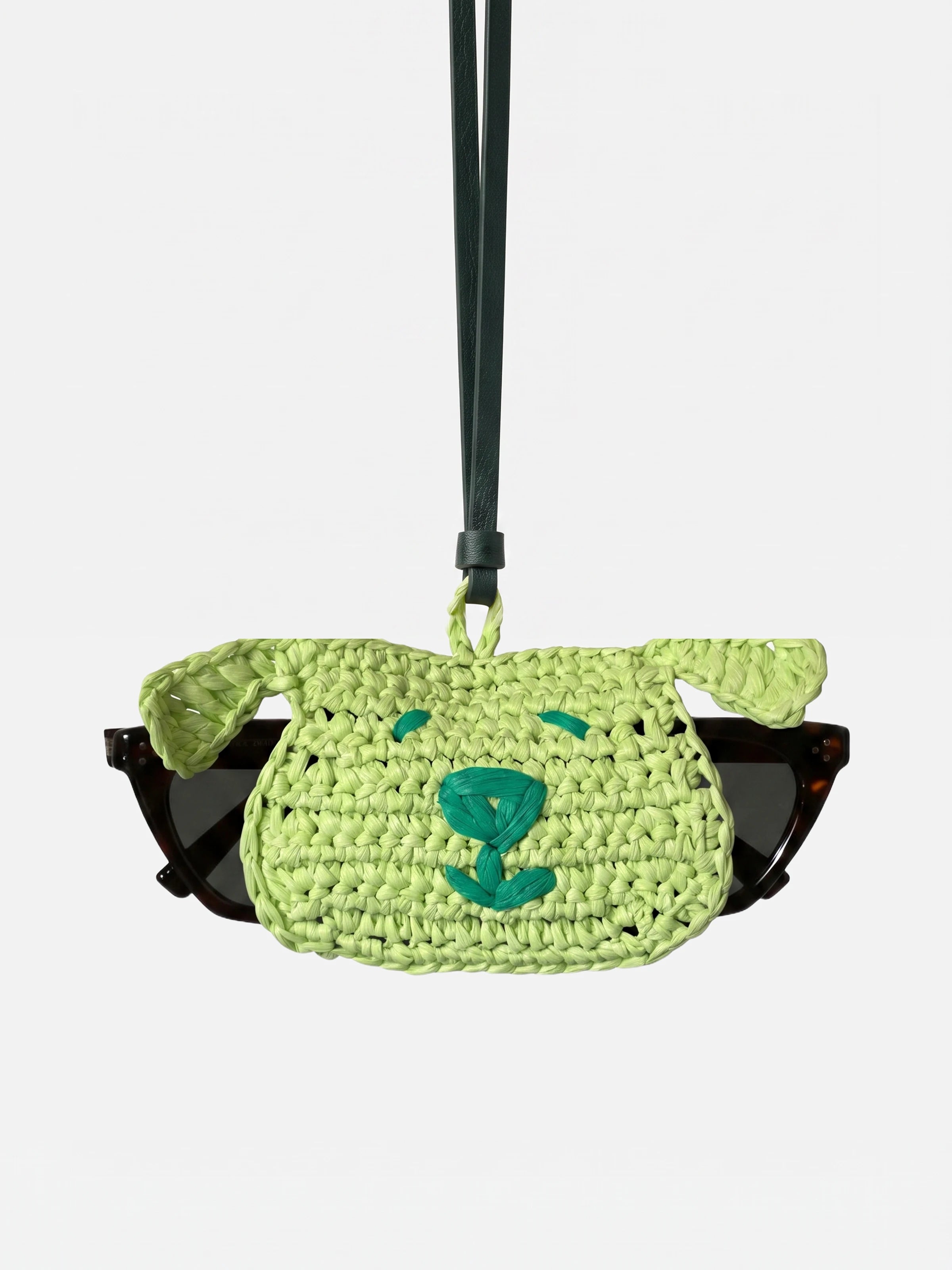 A light green woven sunglasses holder with a dog face design, holding a pair of black sunglasses, suspended from a dark strap against a light gray background.
