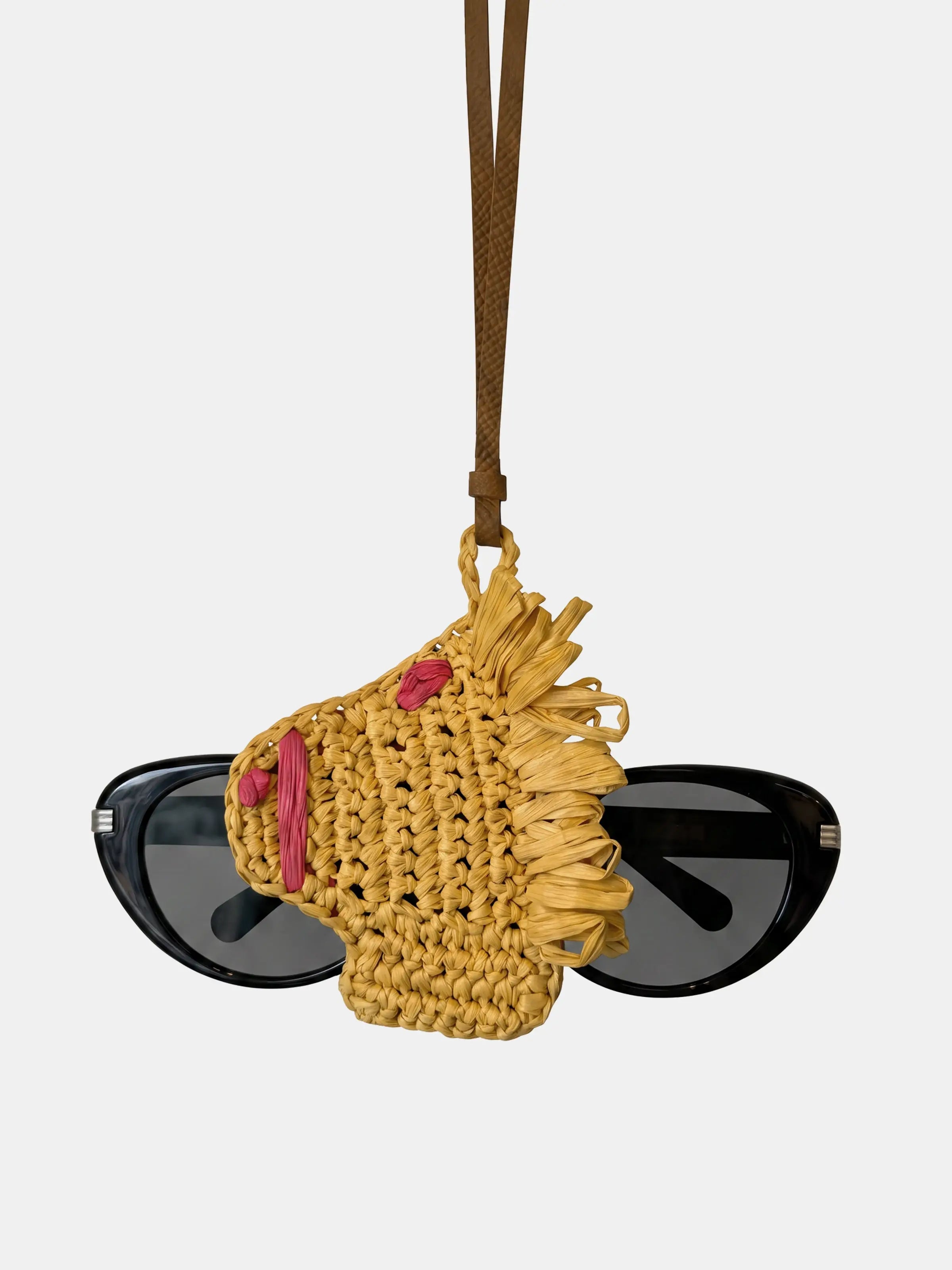 A yellow woven raffia sunglasses pouch with fringe and pink accents, holding a pair of black sunglasses, suspended by a brown strap against a light gray background.