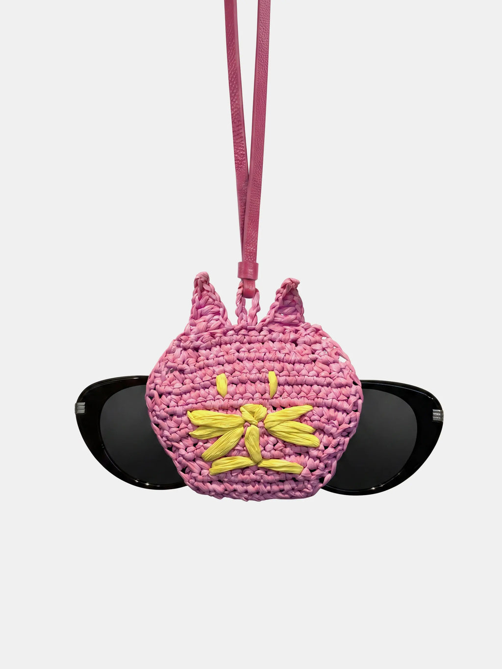 A pink raffia cat sunglasses holder with a yellow embroidered flower on the front, featuring a pink strap and holding a pair of black sunglasses, set against a light gray background.