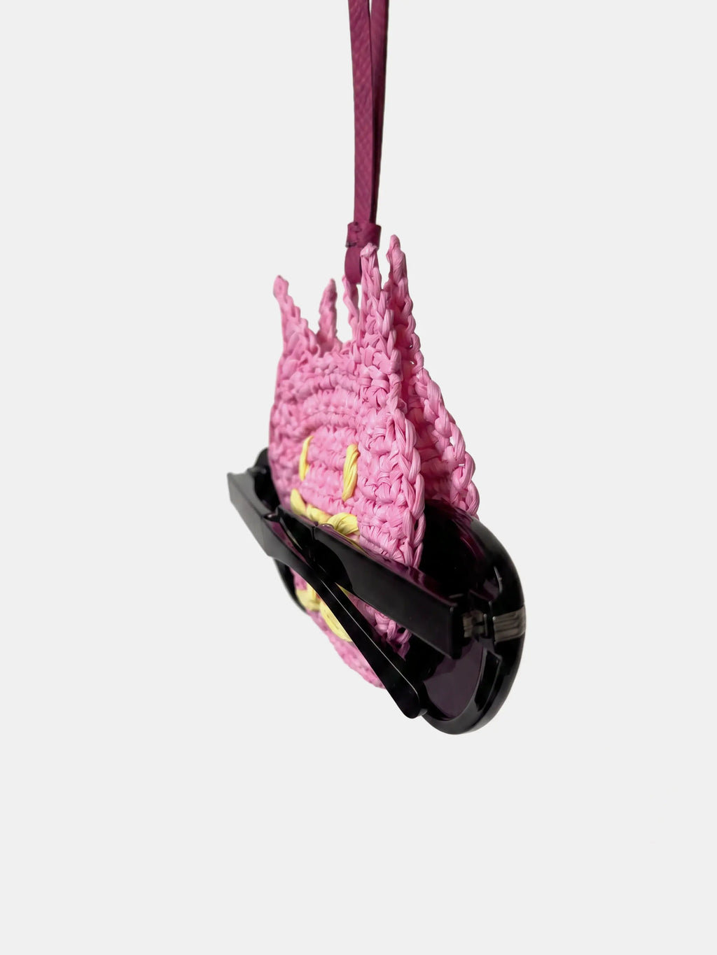 A pink cat raffia charm, holding a pair of black sunglasses, suspended by a dark pink strap against a light gray background.