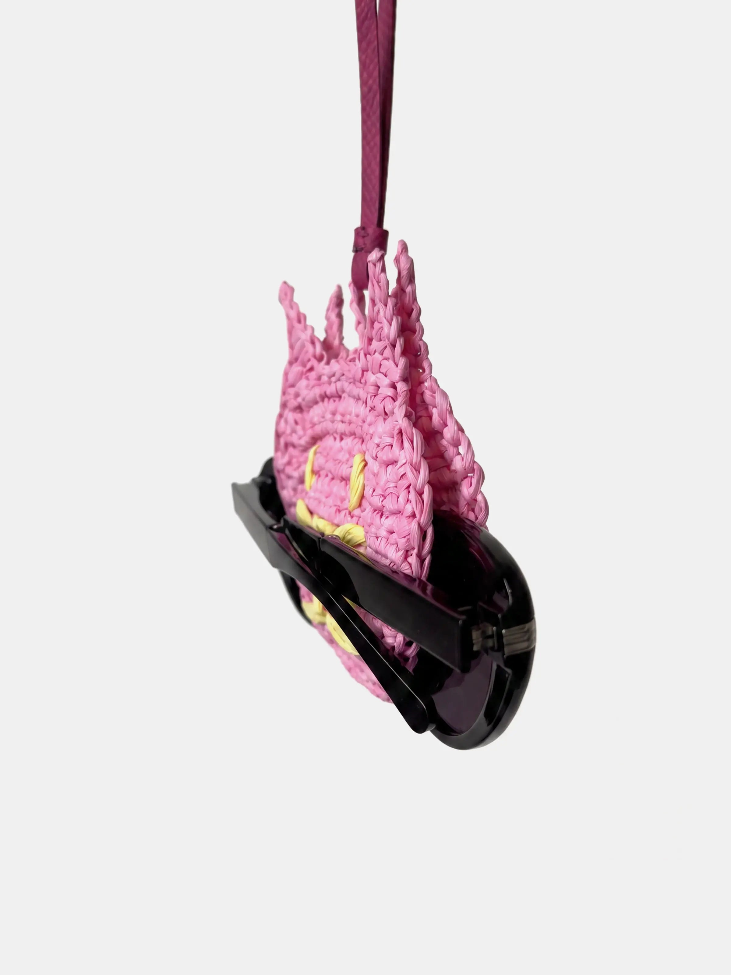 A pink cat raffia charm, holding a pair of black sunglasses, suspended by a dark pink strap against a light gray background.