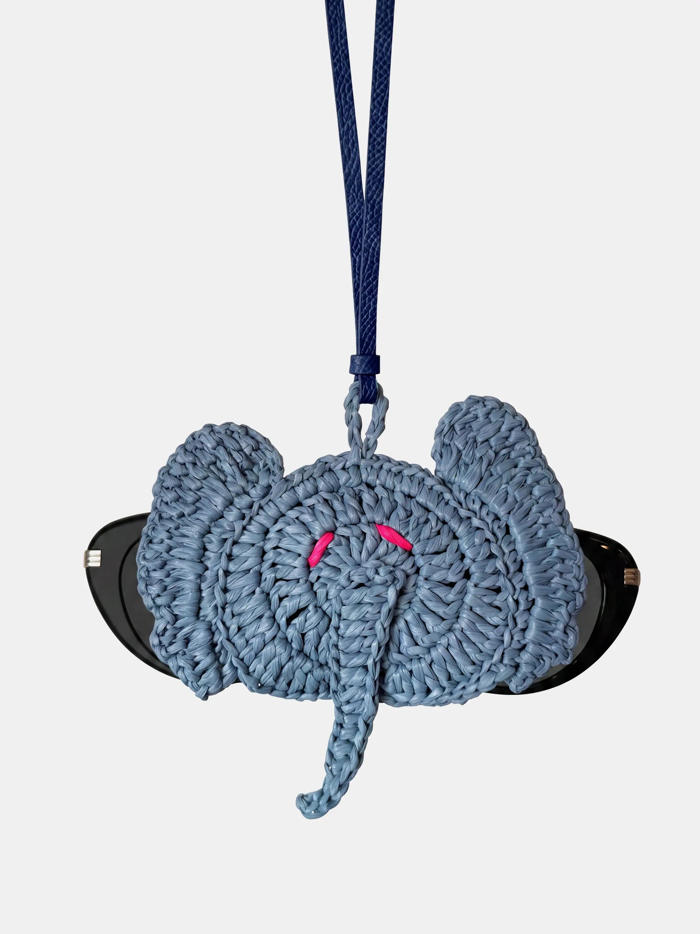 A raffia elephant sunglasses holder in a dusty blue-gray color, featuring a hanging strap and small pink embroidered eyes, shown against a light gray background.