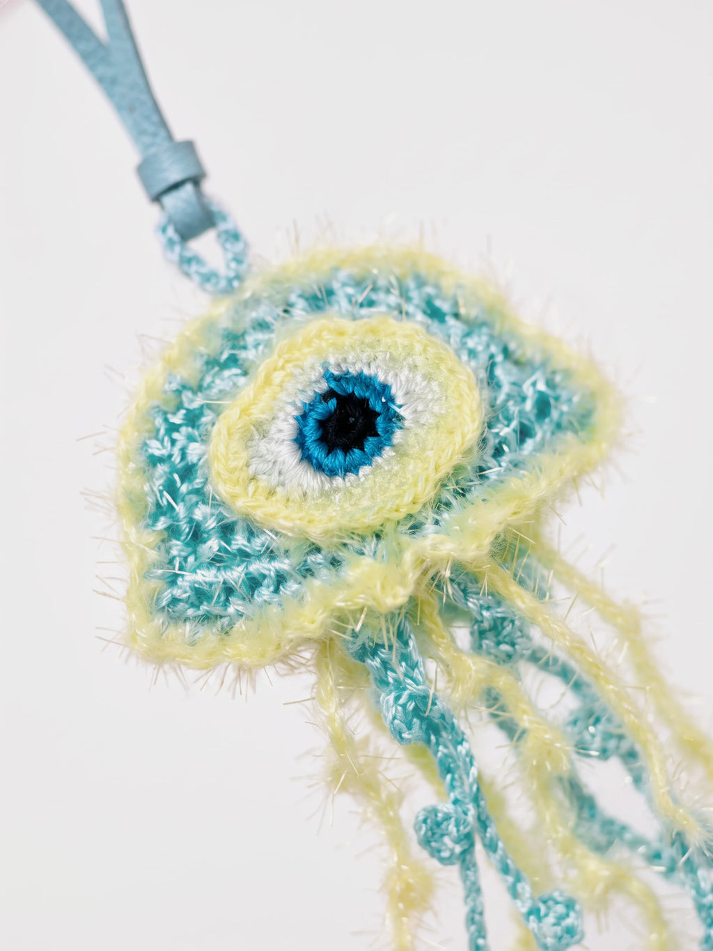 A crocheted or knitted jellyfish raffia charm with a blue strap, featuring a light blue leather strap