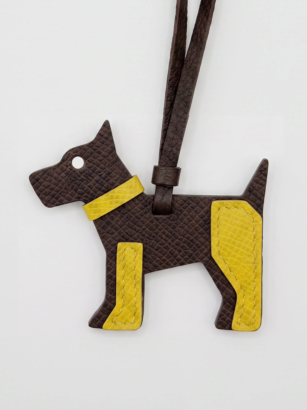 A dog-shaped leather bag charm or luggage tag in dark brown textured leather with bright yellow accents, featuring a matching dark brown strap, shown on a plain light gray background.