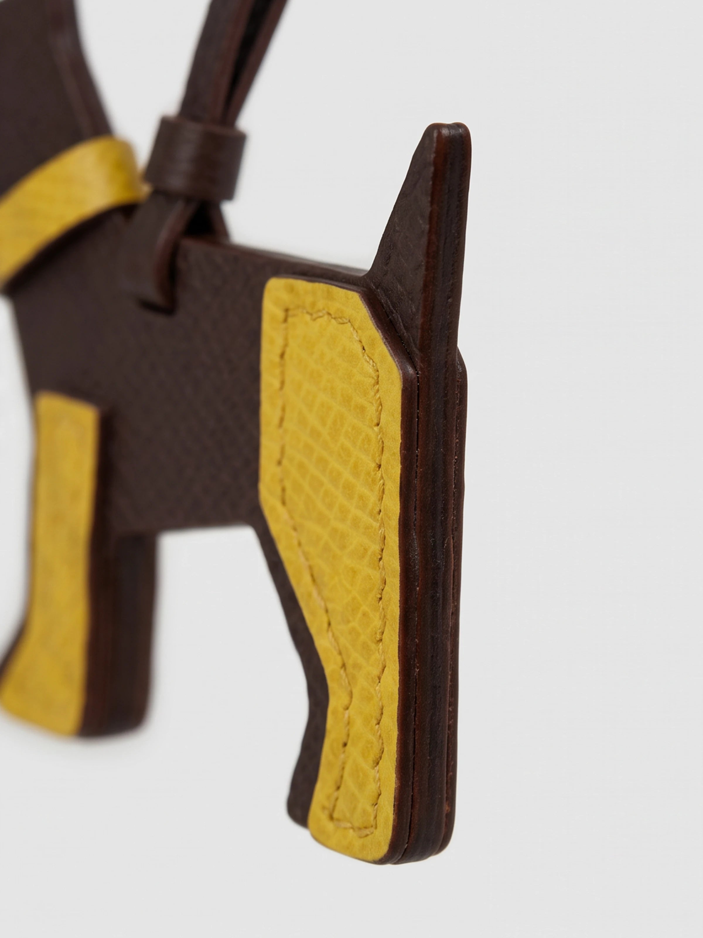 A close-up of a dark brown and mustard yellow leather dog-shaped bag charm or keychain, showing the textured yellow side panels and dark brown edges against a light gray background.