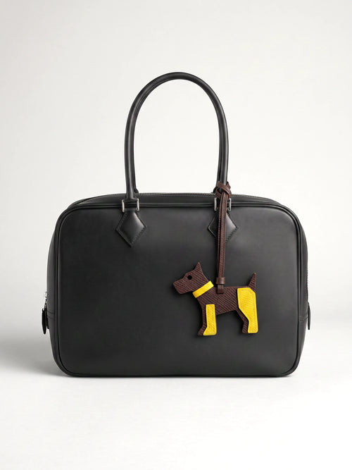 A black leather top-handle satchel bag featuring a decorative dog-shaped charm in brown and yellow leather hanging from the zipper.