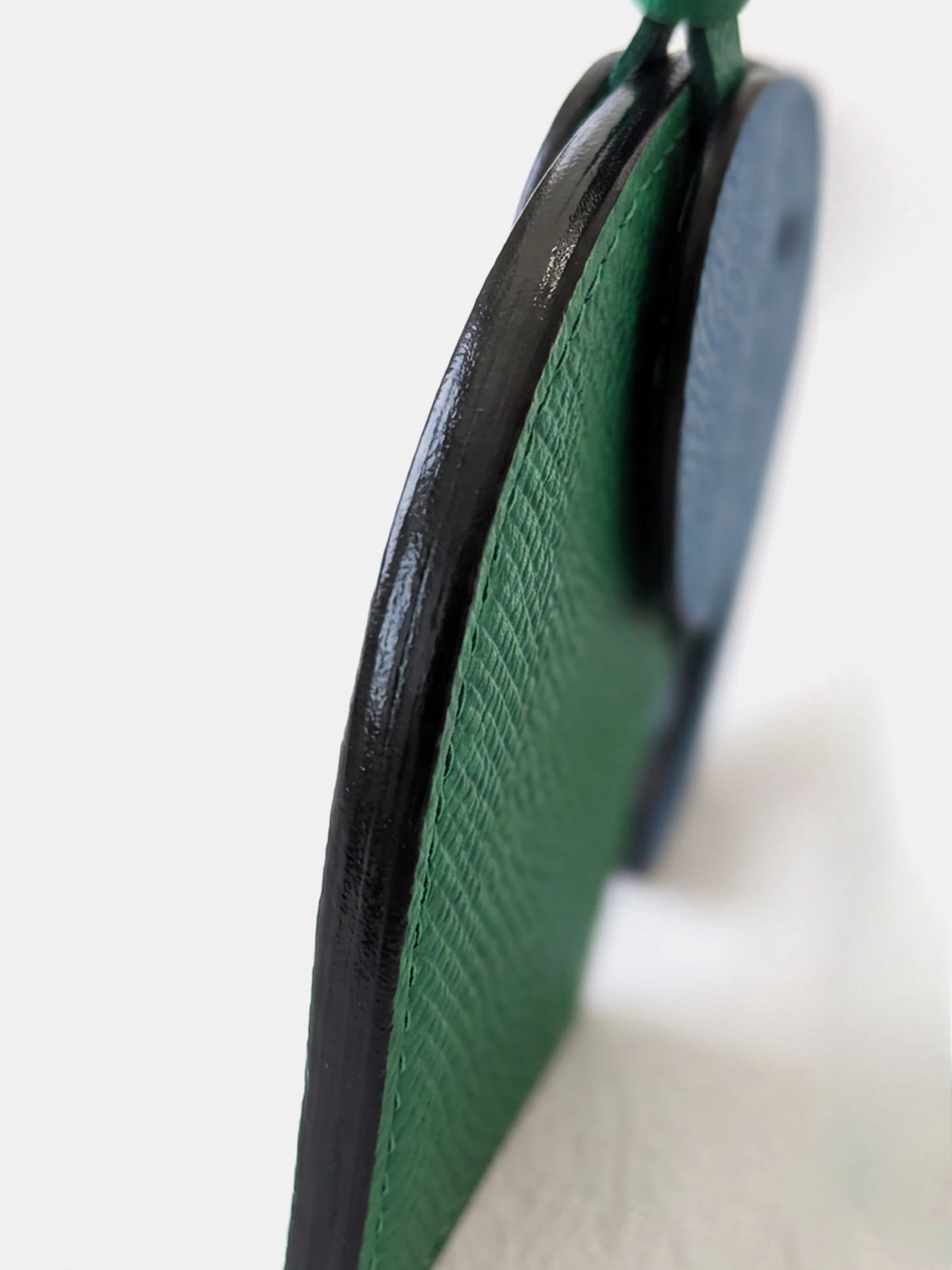 Close-up of a green textured leather accessory with a black edge and a blue circular component, photographed against a clean white background.