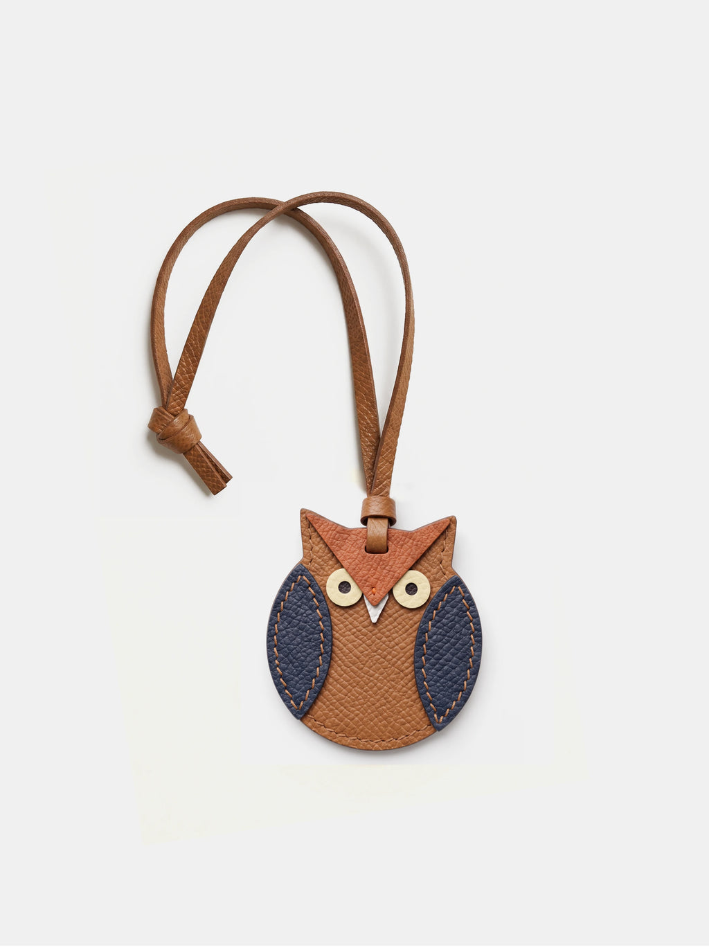 A brown leather owl bag charm with a matching adjustable key strap, featuring navy blue textured wings and cream-colored eyes, set against a light gray background.